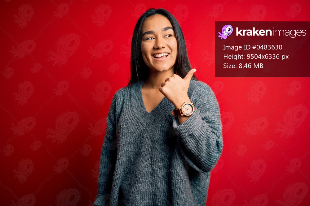 Young beautiful asian woman wearing casual sweater standing over isolated red background smiling with happy face looking and pointing to the side with thumb up.