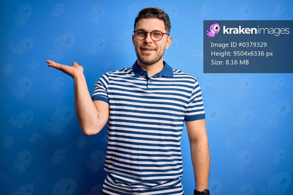 Young man with blue eyes wearing glasses and casual striped t-shirt over blue background smiling cheerful presenting and pointing with palm of hand looking at the camera.