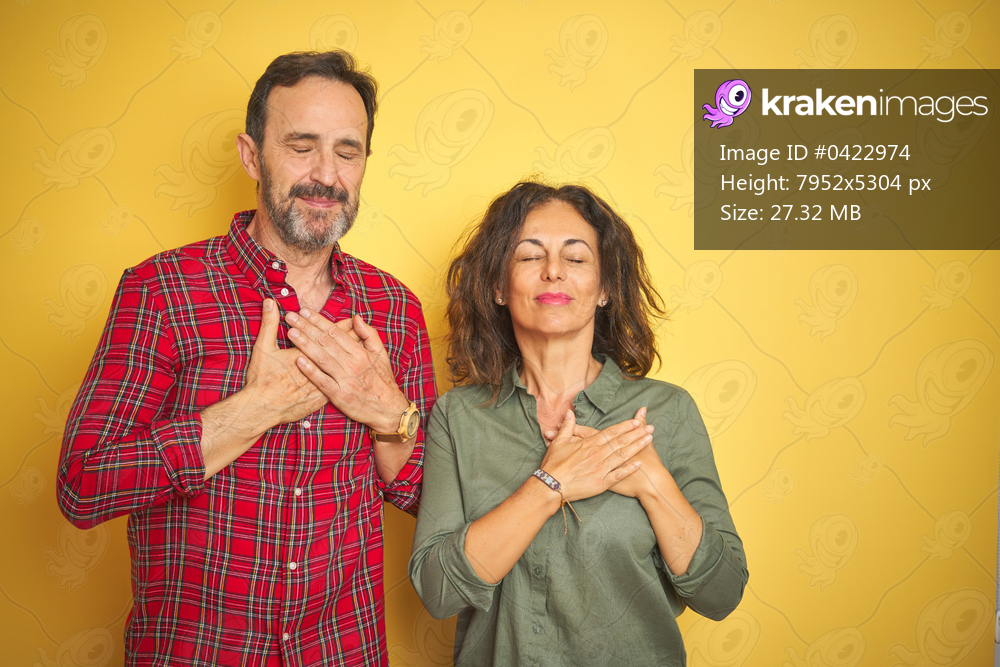 Beautiful middle age couple over isolated yellow background smiling with hands on chest with closed eyes and grateful gesture on face. Health concept.