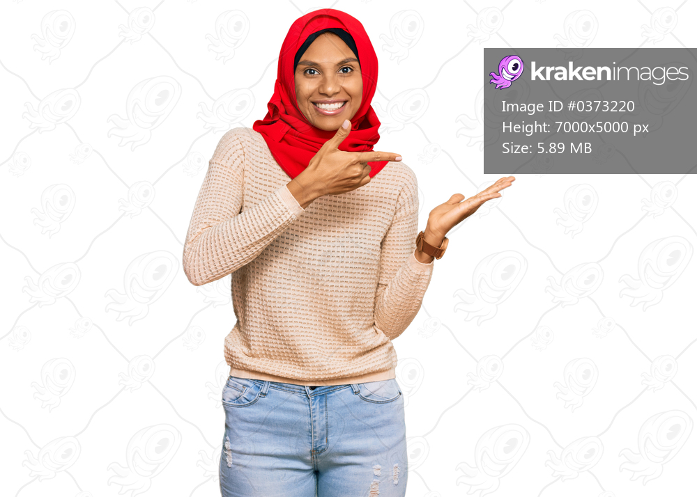 Young african american woman wearing traditional islamic hijab scarf amazed and smiling to the camera while presenting with hand and pointing with finger. 