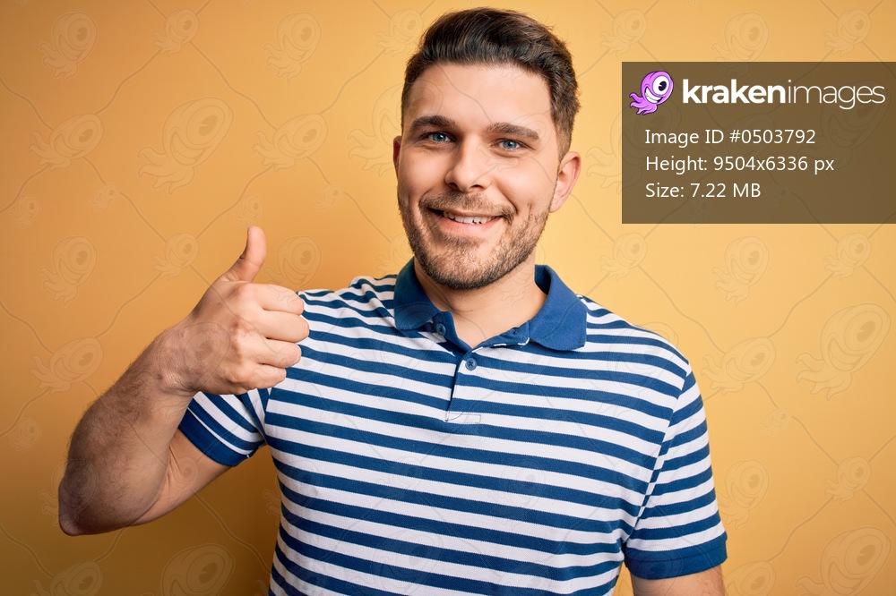 Young man with blue eyes wearing casual striped t-shirt over yellow background doing happy thumbs up gesture with hand. Approving expression looking at the camera showing success.
