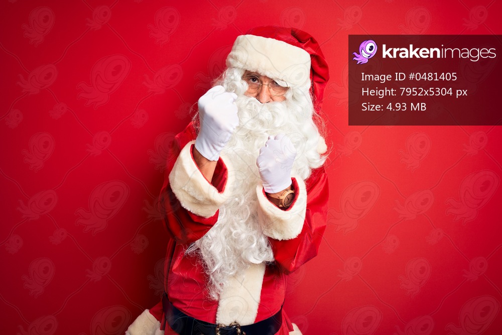 Middle age handsome man wearing Santa costume standing over isolated red background Ready to fight with fist defense gesture, angry and upset face, afraid of problem