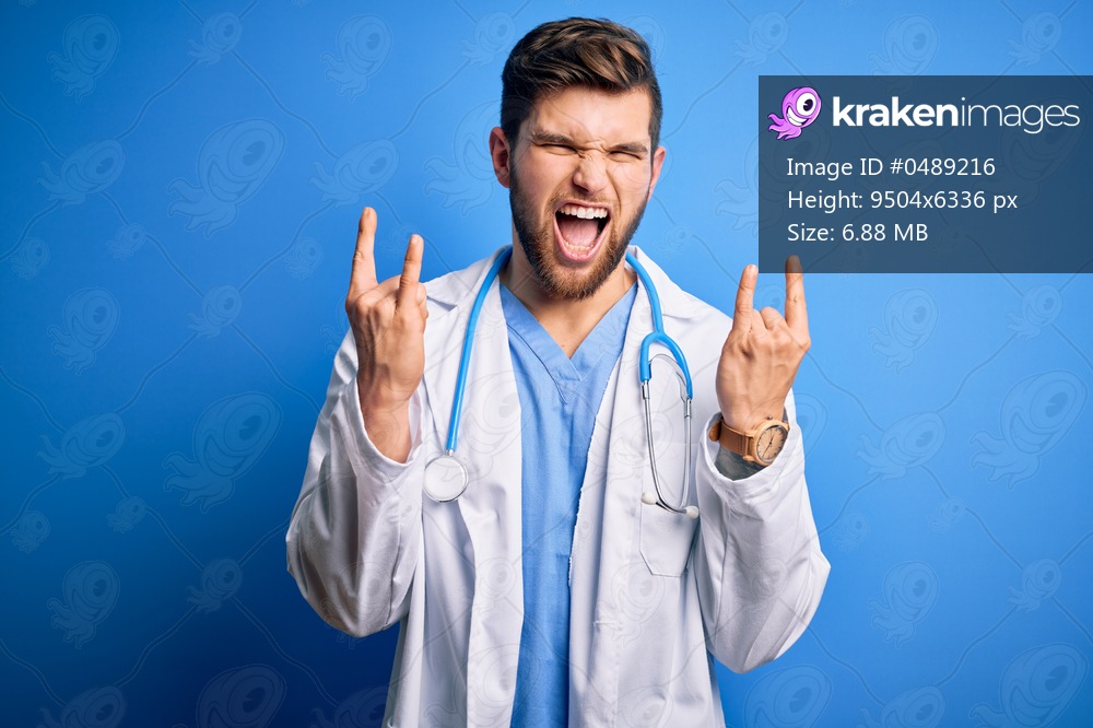 Young blond doctor man with beard and blue eyes wearing white coat and stethoscope shouting with crazy expression doing rock symbol with hands up. Music star. Heavy concept.