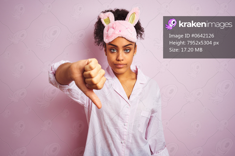 Young african american woman wearing pajama and mask over isolated pink background looking unhappy and angry showing rejection and negative with thumbs down gesture. Bad expression.