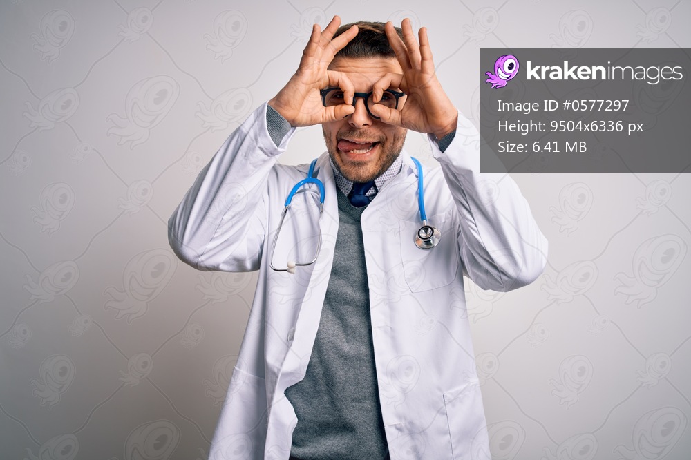 Young doctor man with blue eyes wearing medical coat and stethoscope over isolated background doing ok gesture like binoculars sticking tongue out, eyes looking through fingers. Crazy expression.