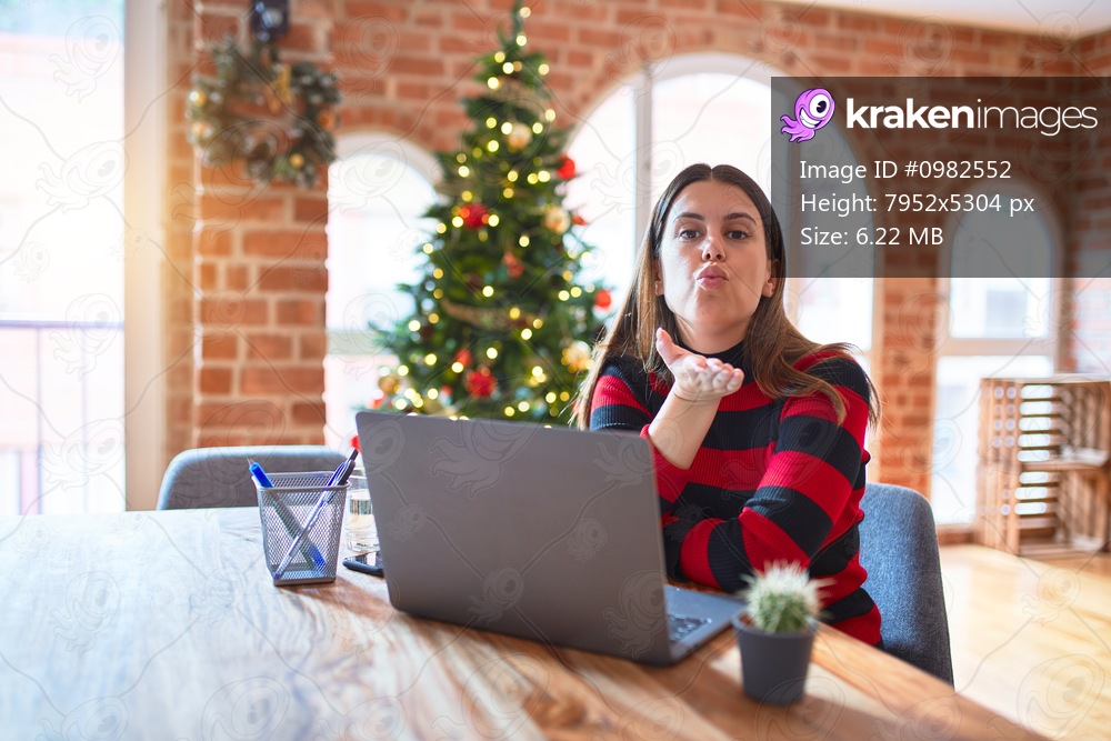 Beautiful woman sitting at the table working with laptop at home around christmas tree looking at the camera blowing a kiss with hand on air being lovely and sexy. Love expression.