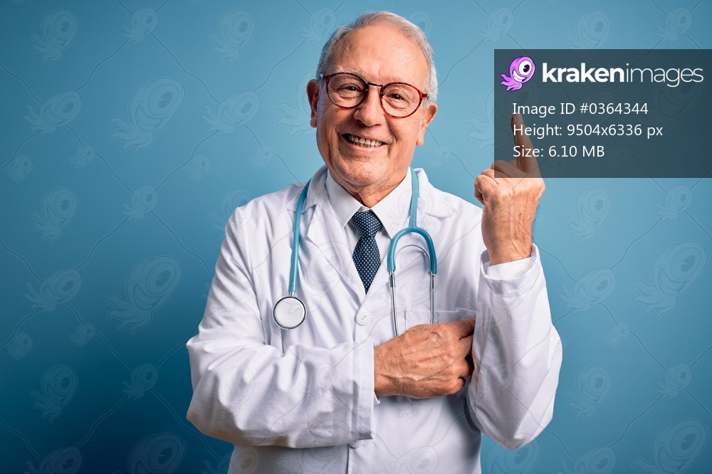 Senior grey haired doctor man wearing stethoscope and medical coat over blue background with a big smile on face, pointing with hand and finger to the side looking at the camera.