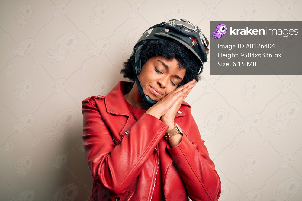 Young African American afro motorcyclist woman with curly hair wearing motorcycle helmet sleeping tired dreaming and posing with hands together while smiling with closed eyes.