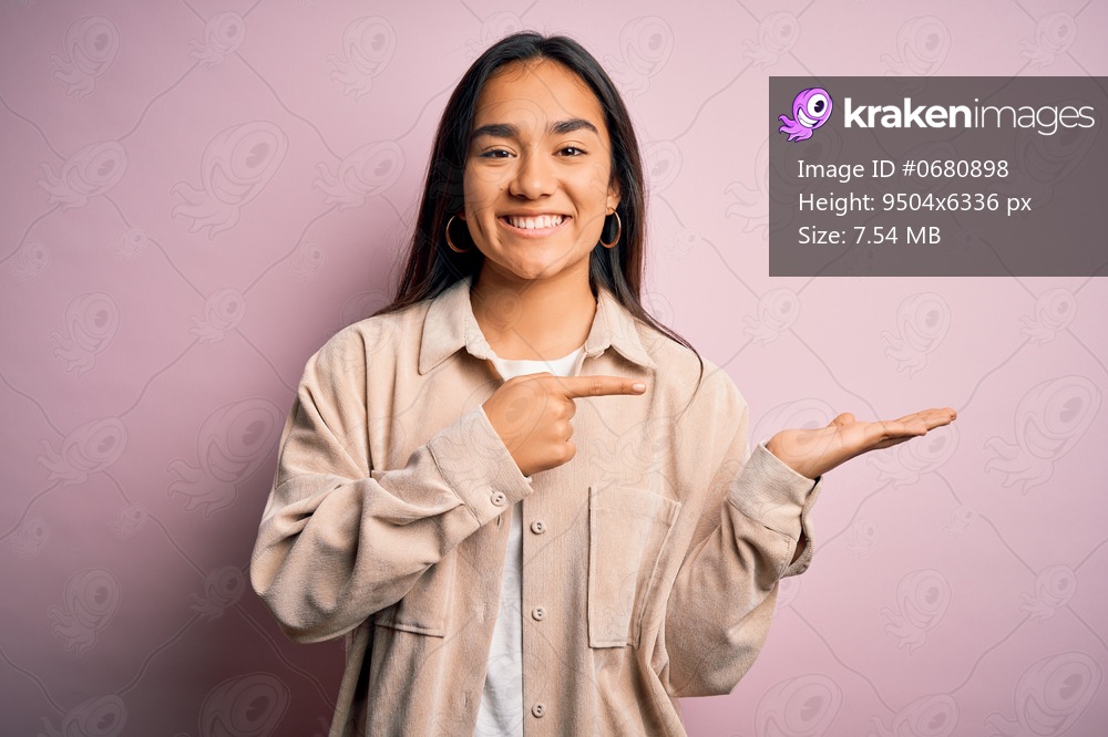 Young beautiful asian woman wearing casual shirt standing over pink background amazed and smiling to the camera while presenting with hand and pointing with finger.