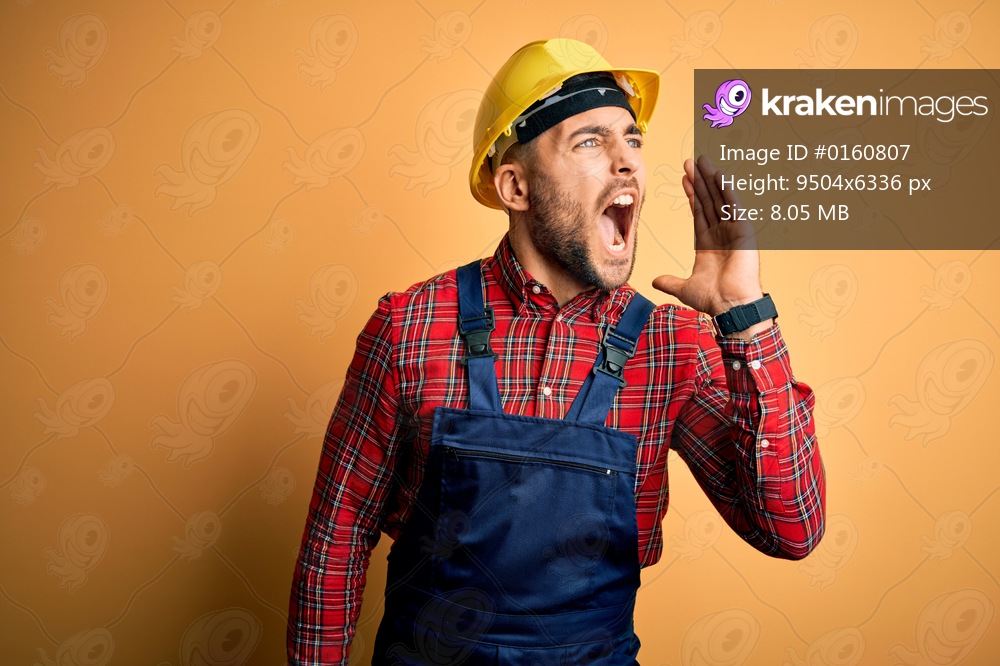 Young builder man wearing construction uniform and safety helmet over yellow isolated background shouting and screaming loud to side with hand on mouth. Communication concept.