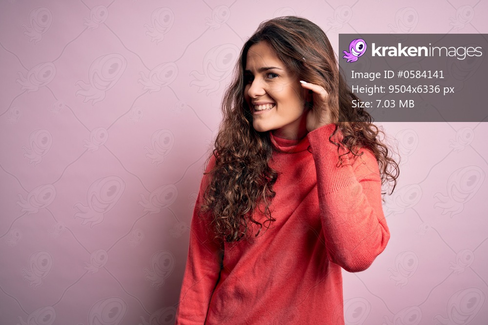 Young beautiful woman with curly hair wearing turtleneck sweater over pink background smiling with hand over ear listening an hearing to rumor or gossip. Deafness concept.