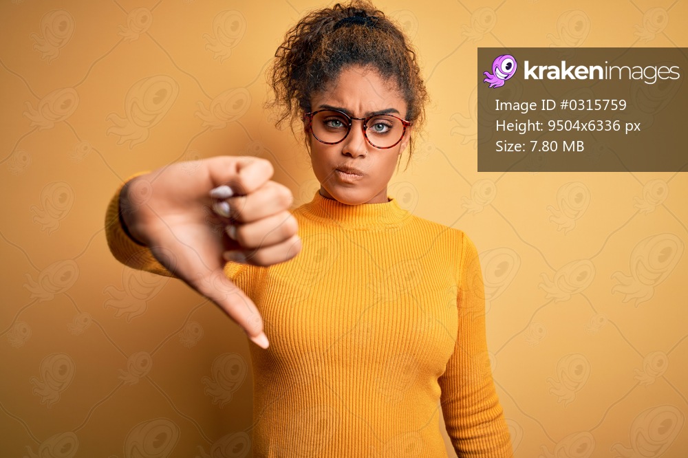 Young beautiful african american girl wearing sweater and glasses over yellow background looking unhappy and angry showing rejection and negative with thumbs down gesture. Bad expression.