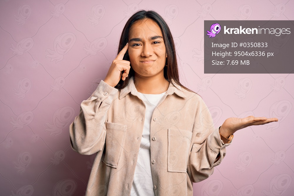 Young beautiful asian woman wearing casual shirt standing over pink background confused and annoyed with open palm showing copy space and pointing finger to forehead. Think about it.