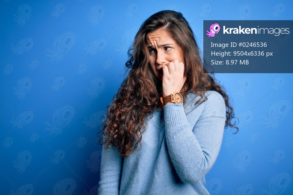 Young beautiful woman with curly hair wearing blue casual sweater over isolated background looking stressed and nervous with hands on mouth biting nails. Anxiety problem.