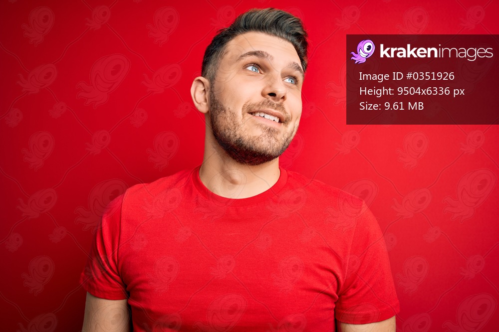 Young man with blue eyes wearing casual t-shirt over red isolated background looking away to side with smile on face, natural expression. Laughing confident.