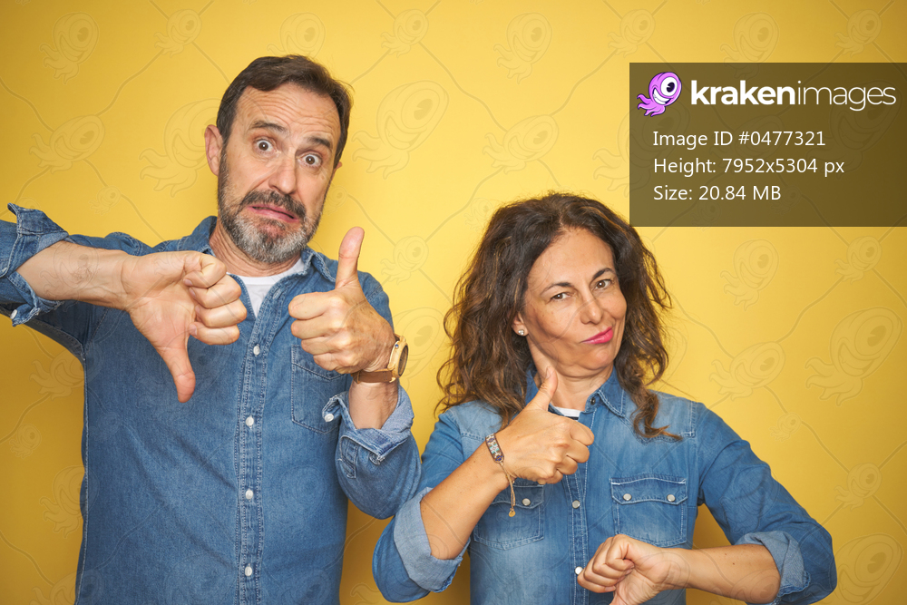 Beautiful middle age couple together standing over isolated yellow background Doing thumbs up and down, disagreement and agreement expression. Crazy conflict
