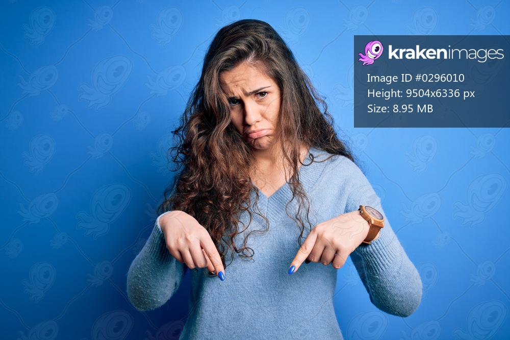 Young beautiful woman with curly hair wearing blue casual sweater over isolated background Pointing down looking sad and upset, indicating direction with fingers, unhappy and depressed.