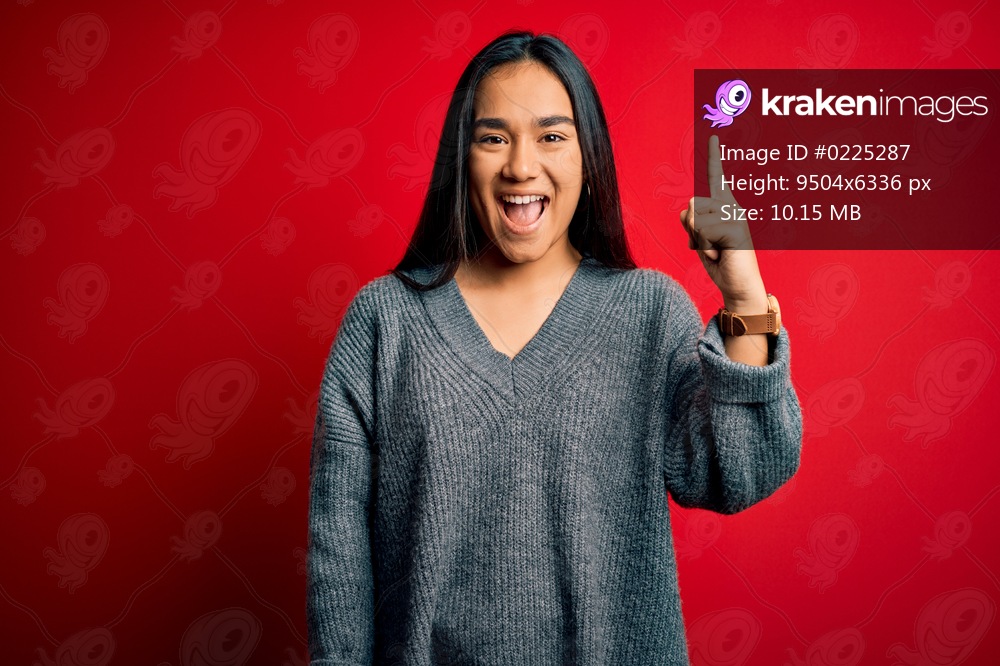 Young beautiful asian woman wearing casual sweater standing over isolated red background pointing finger up with successful idea. Exited and happy. Number one.