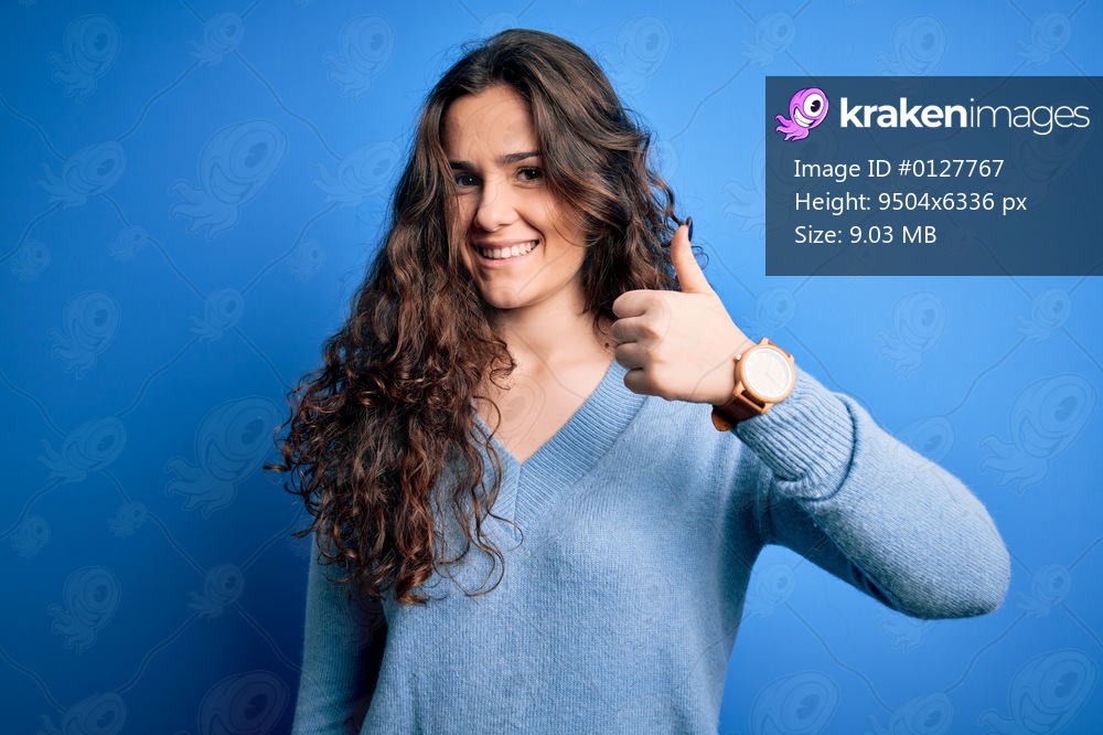 Young beautiful woman with curly hair wearing blue casual sweater over isolated background doing happy thumbs up gesture with hand. Approving expression looking at the camera showing success.