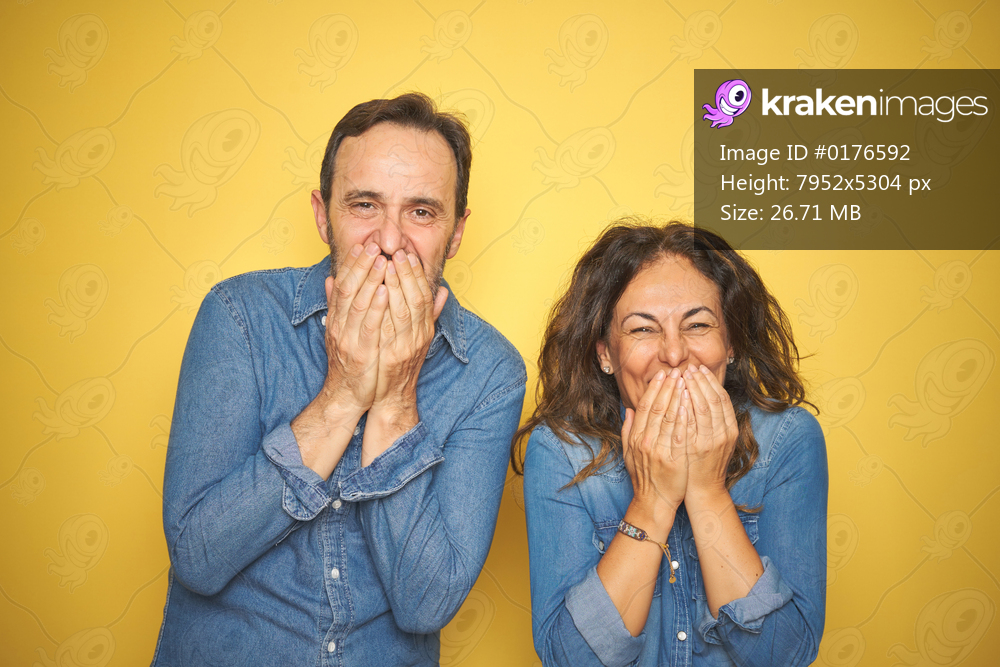 Beautiful middle age couple together standing over isolated yellow background laughing and embarrassed giggle covering mouth with hands, gossip and scandal concept