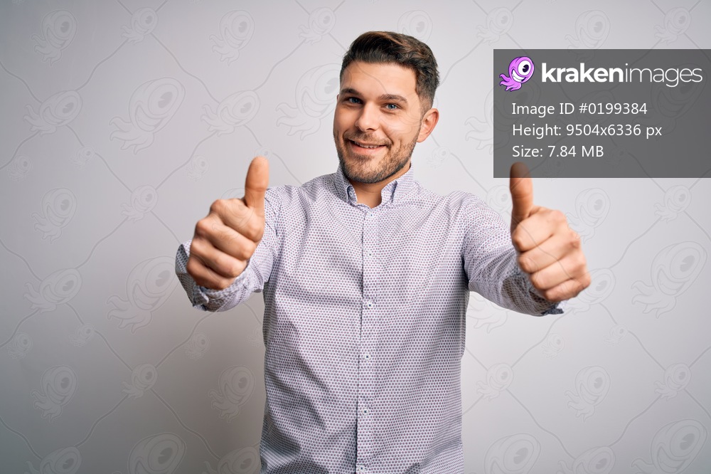 Young business man with blue eyes standing over isolated background approving doing positive gesture with hand, thumbs up smiling and happy for success. Winner gesture.