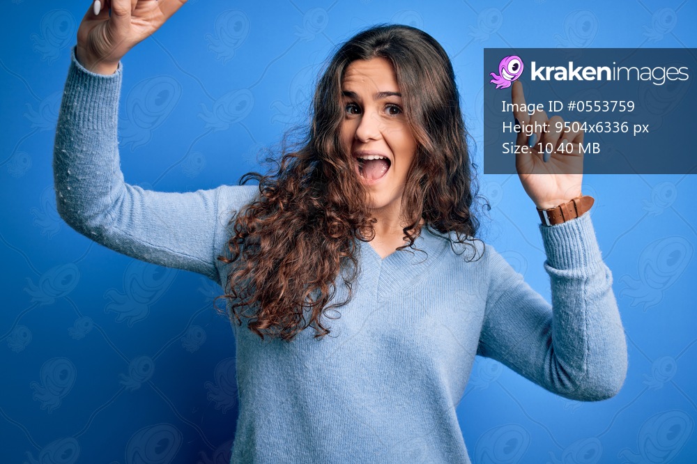 Young beautiful woman with curly hair wearing blue casual sweater over isolated background smiling amazed and surprised and pointing up with fingers and raised arms.