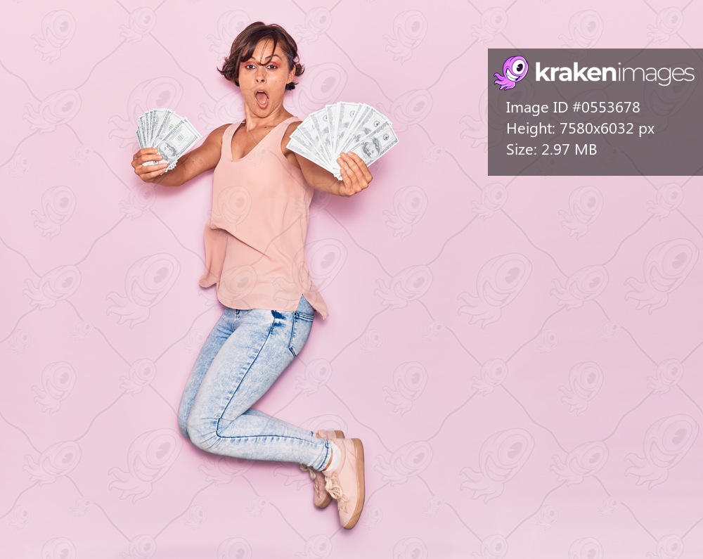 Young beautiful woman surprised holding dollars banknotes . Jumping with open mouth over isolated pink background.