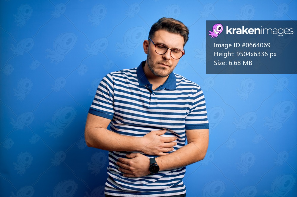 Young man with blue eyes wearing glasses and casual striped t-shirt over blue background with hand on stomach because indigestion, painful illness feeling unwell. Ache concept.