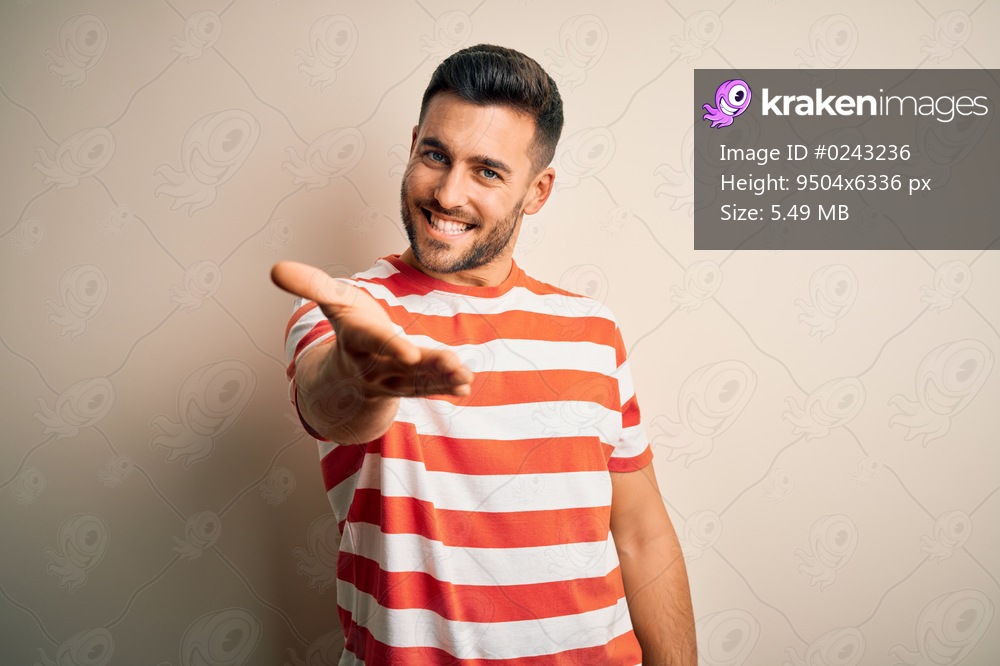 Young handsome man wearing casual striped t-shirt standing over isolated white background smiling cheerful offering palm hand giving assistance and acceptance.