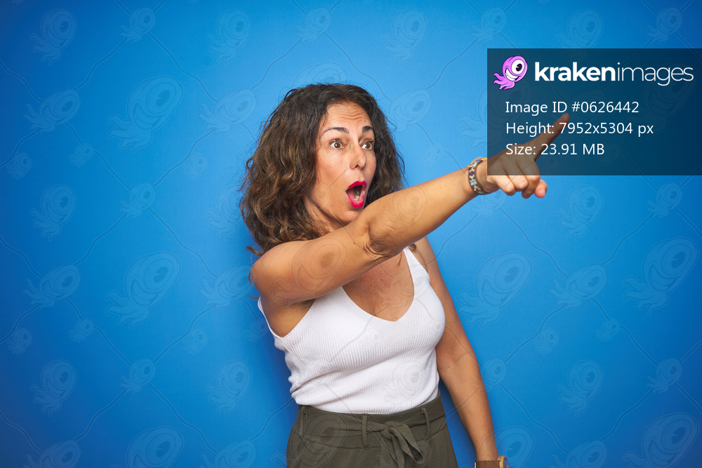 Middle age senior woman with curly hair standing over blue isolated background Pointing with finger surprised ahead, open mouth amazed expression, something on the front