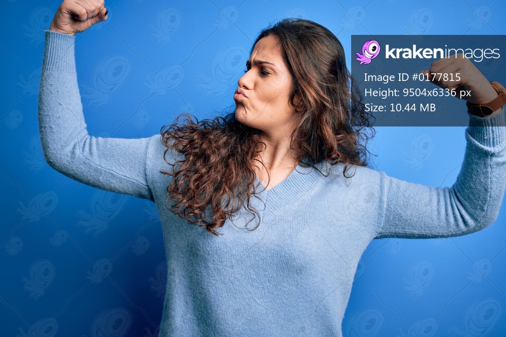 Young beautiful woman with curly hair wearing blue casual sweater over isolated background showing arms muscles smiling proud. Fitness concept.