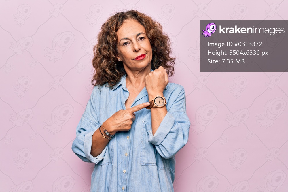 Middle age beautiful woman wearing casual denim shirt standing over pink background In hurry pointing to watch time, impatience, looking at the camera with relaxed expression