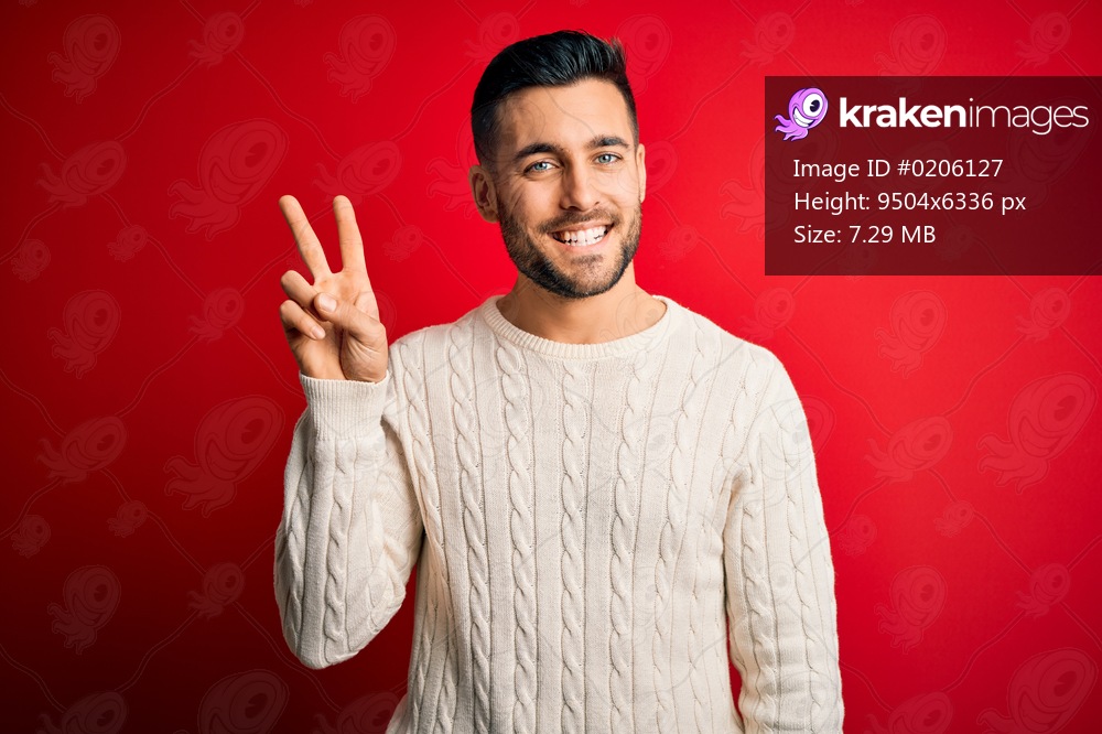 Young handsome man wearing casual white sweater standing over isolated red background showing and pointing up with fingers number two while smiling confident and happy.