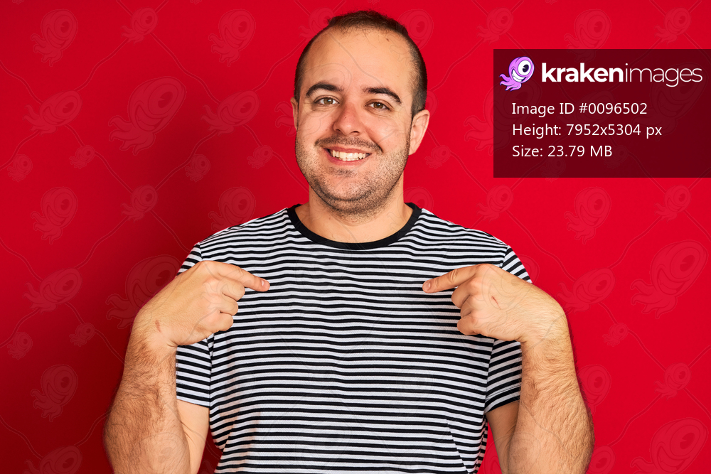 Young man wearing striped navy t-shirt standing over isolated red background looking confident with smile on face, pointing oneself with fingers proud and happy.
