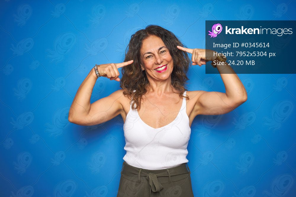 Middle age senior woman with curly hair standing over blue isolated background smiling pointing to head with both hands finger, great idea or thought, good memory