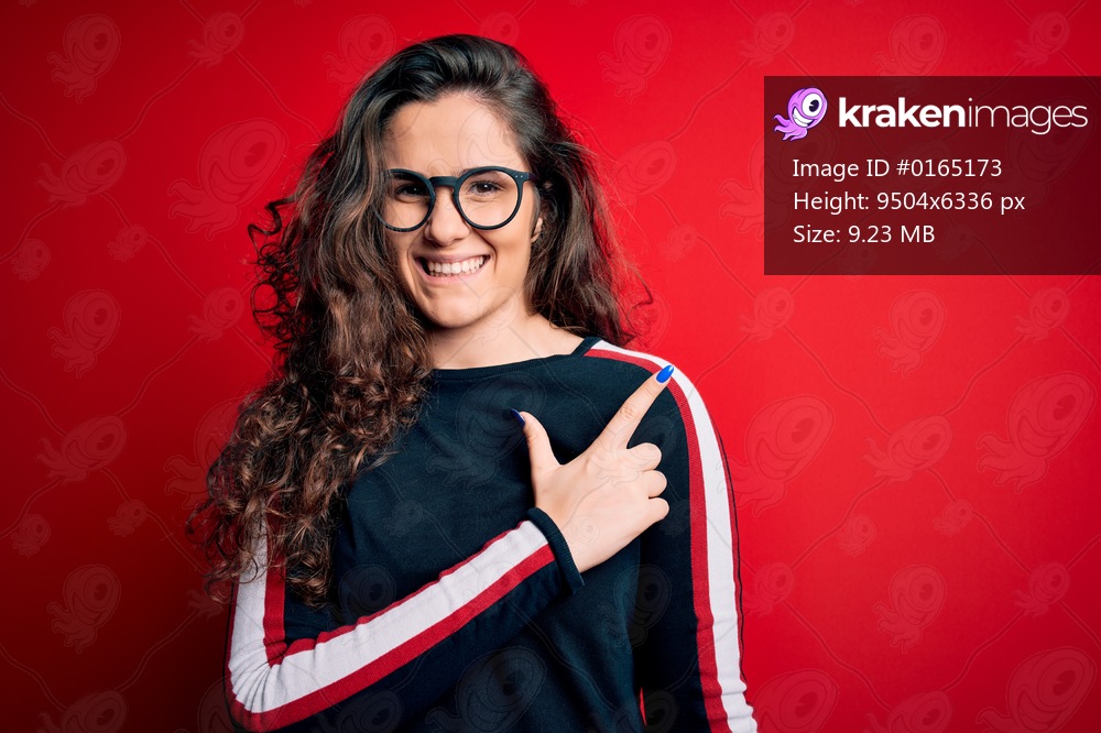 Young beautiful woman with curly hair wearing sweater and glasses over red background cheerful with a smile of face pointing with hand and finger up to the side with happy and natural expression on face