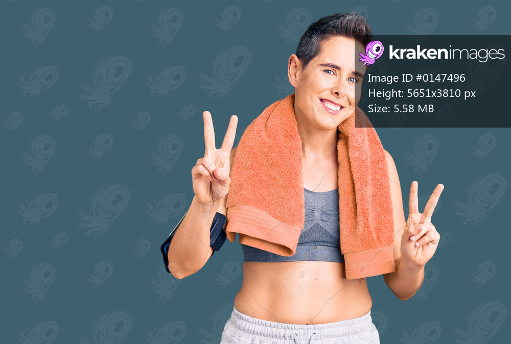 Young woman with short hair wearing sportswear and towel using smartphone smiling looking to the camera showing fingers doing victory sign. number two. 