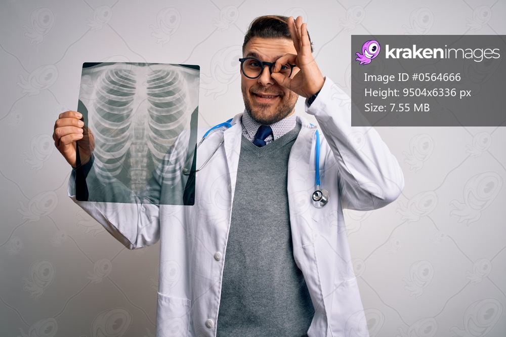 Young doctor man wearing medical coat looking at chest scan radiography over isolated background with happy face smiling doing ok sign with hand on eye looking through fingers