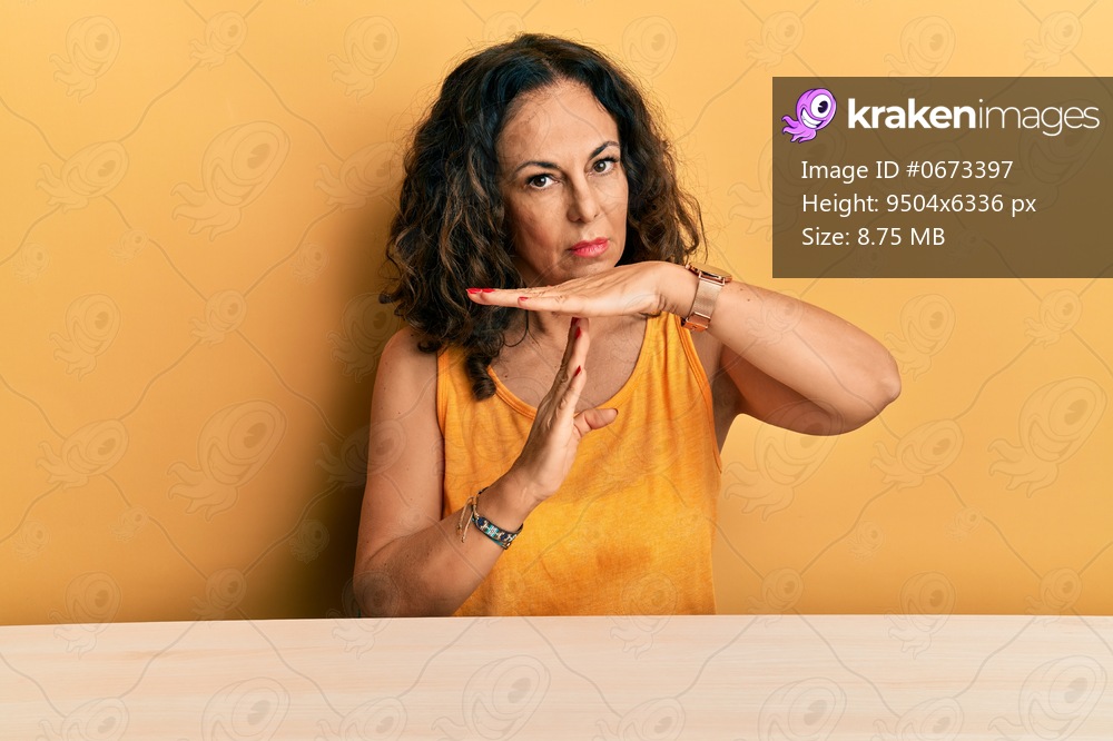 Beautiful middle age woman wearing casual clothes sitting on the table doing time out gesture with hands, frustrated and serious face 
