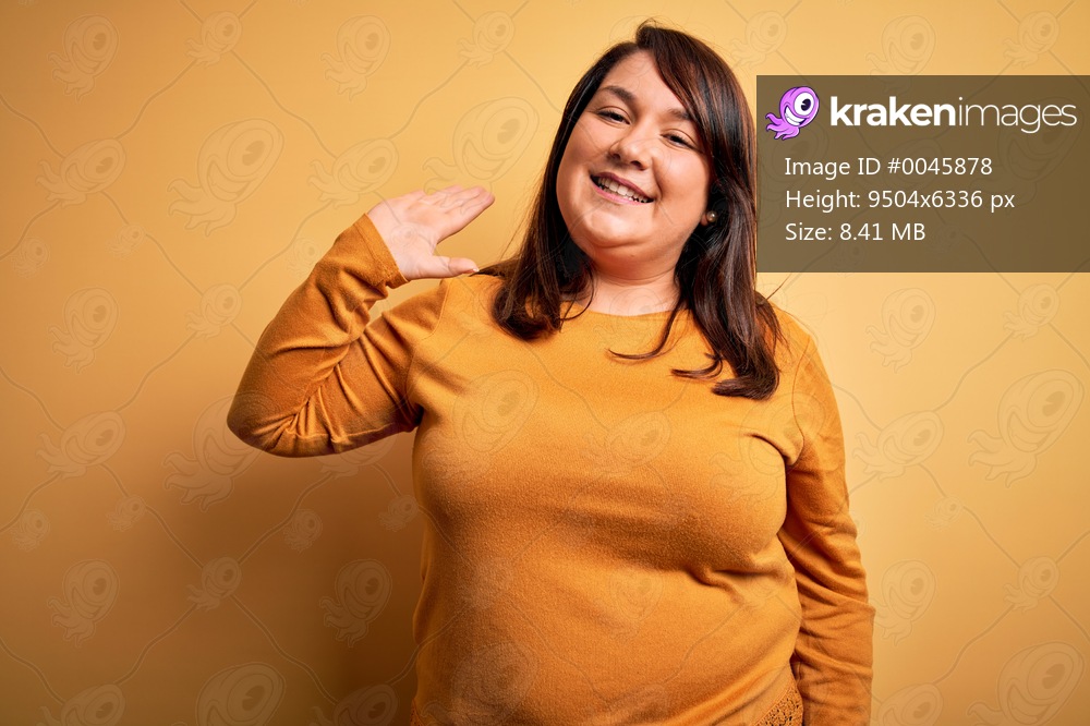 Beautiful brunette plus size woman wearing casual sweater over isolated yellow background Waiving saying hello happy and smiling, friendly welcome gesture
