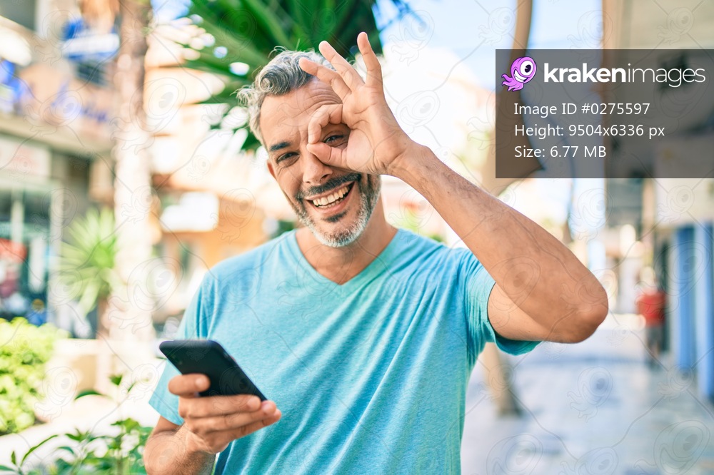 Middle age grey-haired man using smartphone at street of city smiling happy doing ok sign with hand on eye looking through fingers 