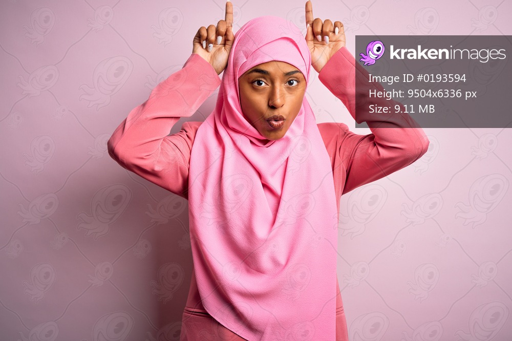 Young African American afro woman wearing muslim hijab over isolated pink background doing funny gesture with finger over head as bull horns
