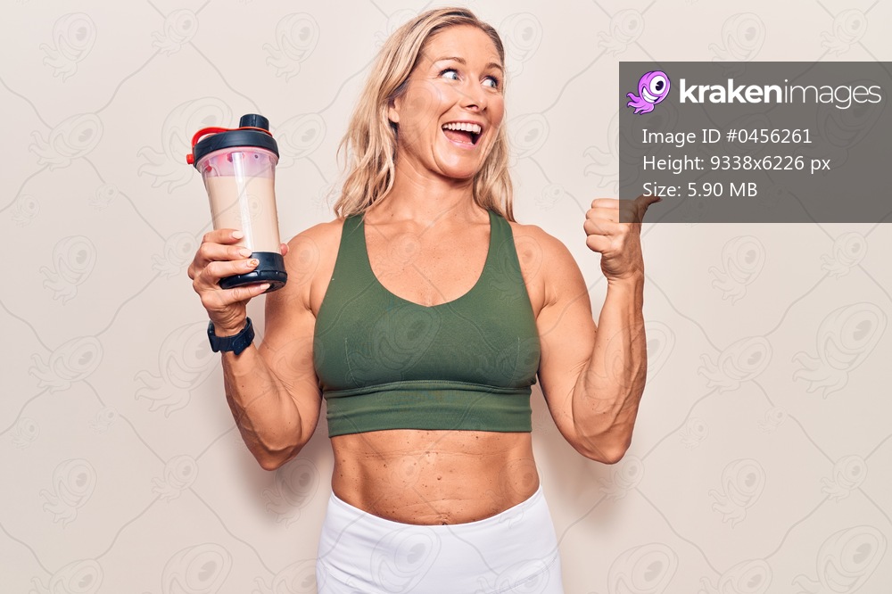 Middle age caucasian blonde woman wearing sport clothes drinking a protein shake pointing thumb up to the side smiling happy with open mouth 