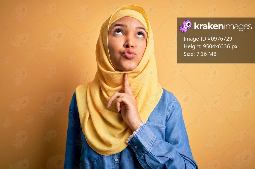 Young beautiful woman with curly hair wearing arab traditional hijab over yellow background Thinking concentrated about doubt with finger on chin and looking up wondering