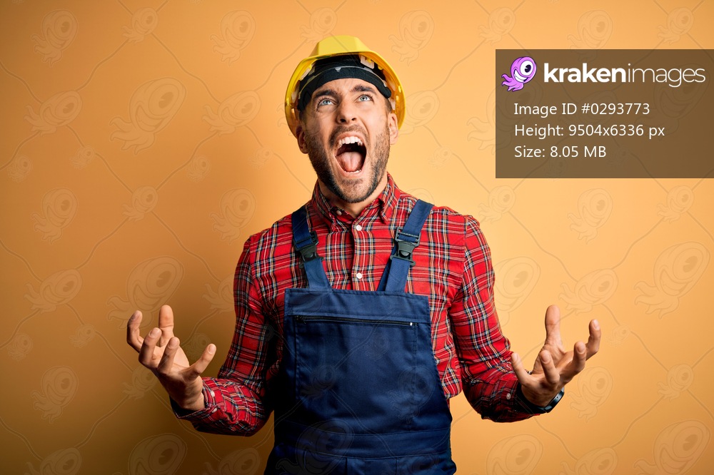 Young builder man wearing construction uniform and safety helmet over yellow isolated background crazy and mad shouting and yelling with aggressive expression and arms raised. Frustration concept.