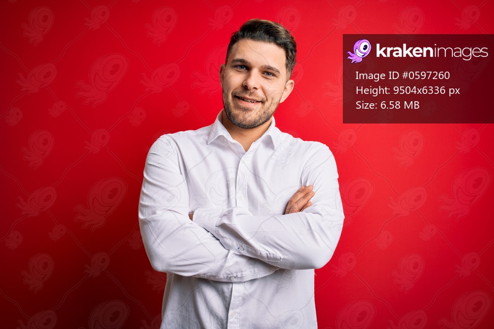 Young business man with blue eyes wearing elegant shirt standing over red isolated background happy face smiling with crossed arms looking at the camera. Positive person.