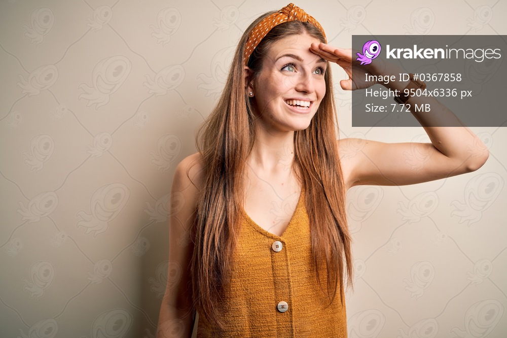 Young beautiful redhead woman wearing casual t-shirt and diadem over yellow background very happy and smiling looking far away with hand over head. Searching concept.