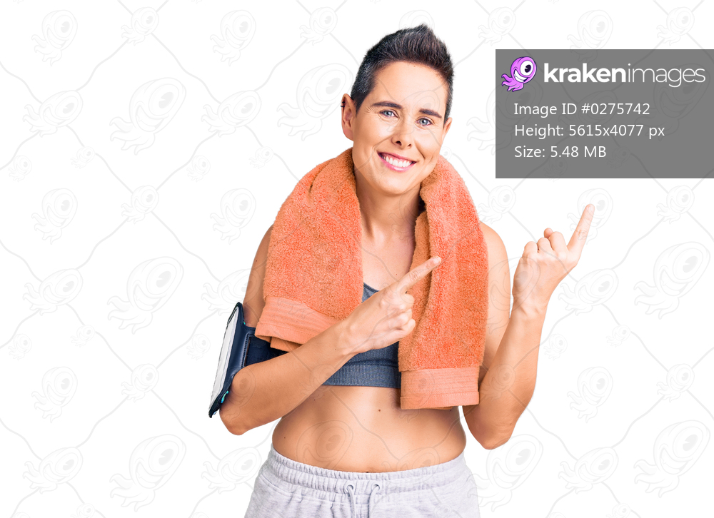Young woman with short hair wearing sportswear and towel using smartphone smiling and looking at the camera pointing with two hands and fingers to the side. 