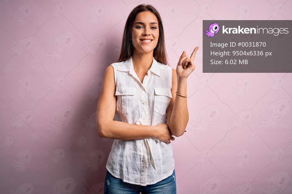 Young beautiful brunette woman wearing casual shirt standing over pink background with a big smile on face, pointing with hand and finger to the side looking at the camera.