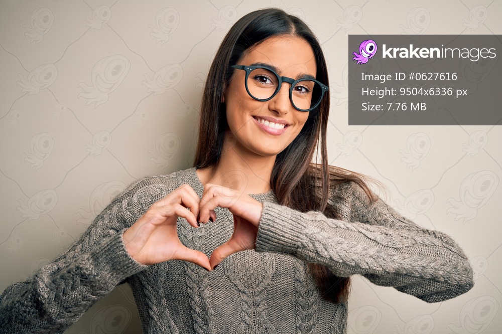 Young beautiful brunette woman wearing casual sweater and glasses over white background smiling in love showing heart symbol and shape with hands. Romantic concept.